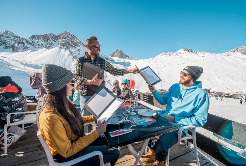 A couple sit at a restaurant on the outside terrace under the sun, as a waiter hands them menus