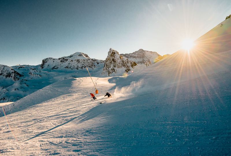 two skiers ski fast down a piste, at what looks like afternoon last light with the sun about to disappear behind the mountain