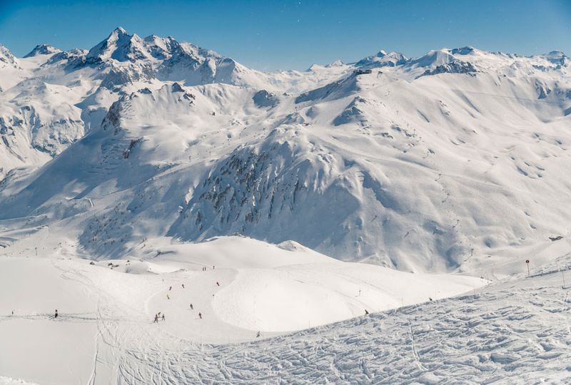a very white, snowy mountainscape with a piste in the mid-distance dotted with tiny silhouettes of skiers
